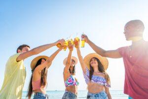 A group of young friends in summer clothes and straw hats toasting with beer bottles on a sunny beach.