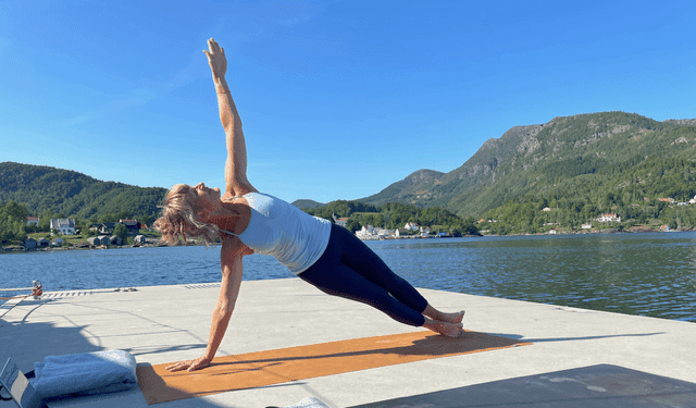 A woman practicing a side plank yoga pose on a wooden jetty with mountains and calm sea in the background